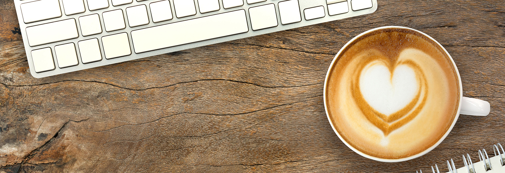Latte with heart-shaped foam on desk with keyboard and notebook