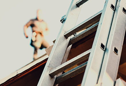 Ladder leaning on a building, out of focus worker walks on roof in background