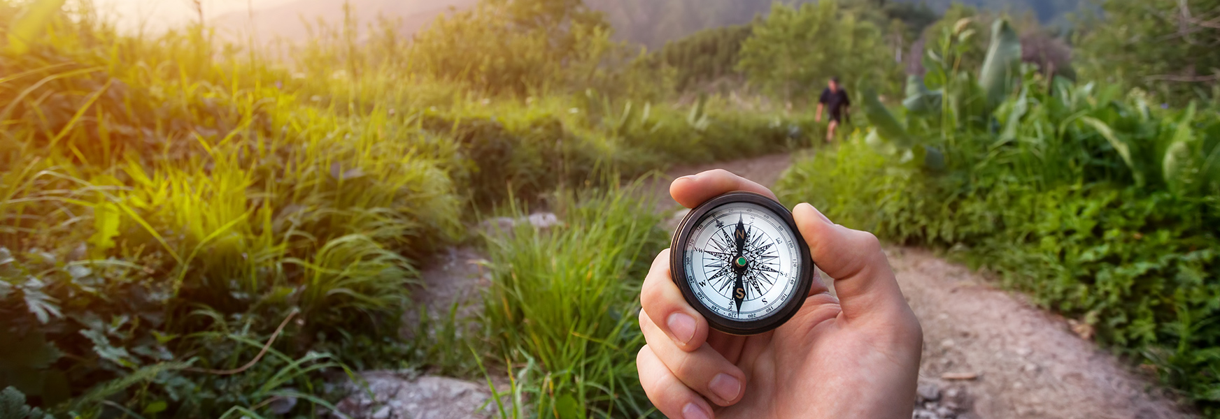 Hand holding a compass with out of focus mountains, tropical vegetation, and hiking partner in background