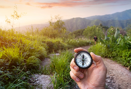 Hand holding a compass with out of focus mountains, tropical vegetation, and hiking partner in background