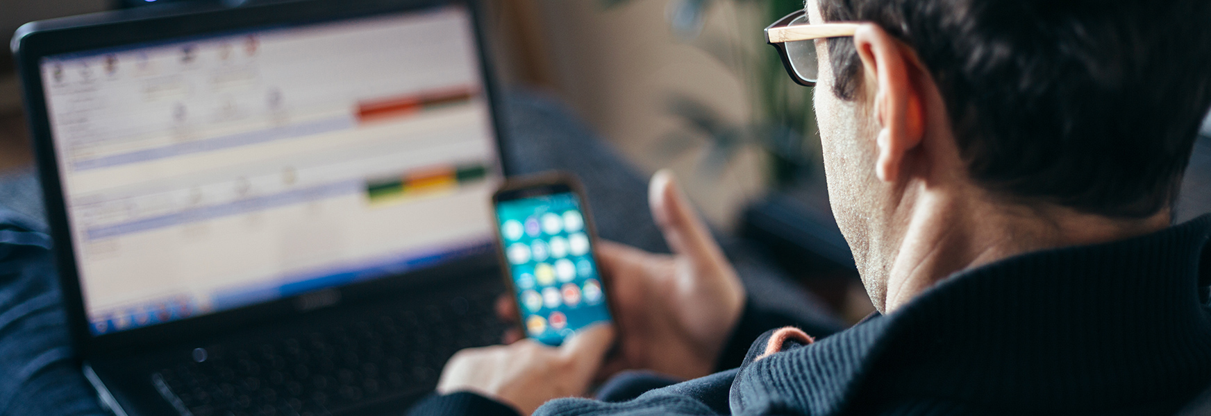 Man checks his cellphone while working at home with laptop on his lap