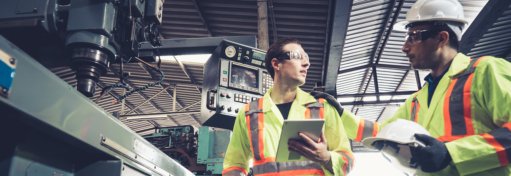 Two men in factory setting, one helmet-wearing man offers a helmet to the other man, who is not wearing one.