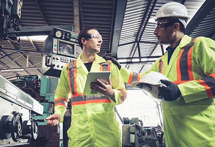 Two men in factory setting, one helmet-wearing man offers a helmet to the other man, who is not wearing one.